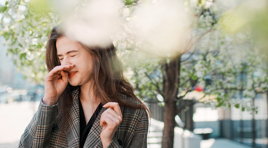young woman holds a sneeze while outside on a tree-lined city street