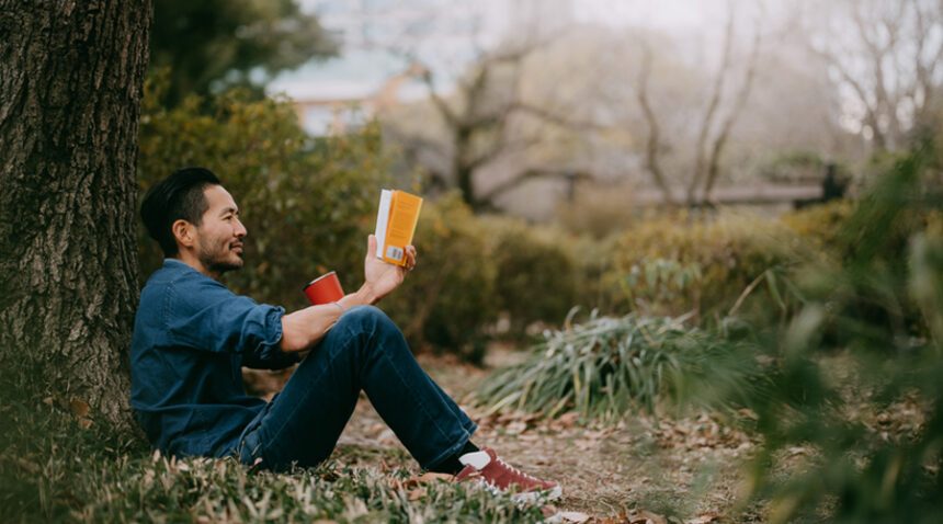Man sitting in a park, reading a book and holding a drink mug.