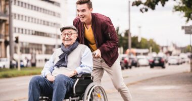 Young man pushes an elderly man in a wheelchair. Both are smiling.