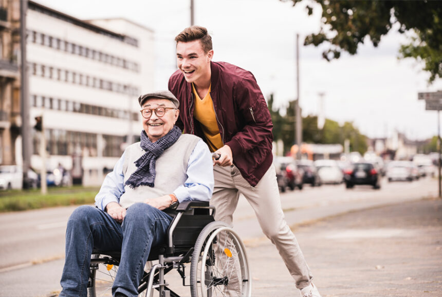 Young man pushes an elderly man in a wheelchair. Both are smiling.