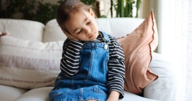 little girl sits on a couch, leans head to the side while she clutches her ear