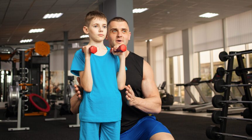 child holds small weights while dad coaches him in gym envrionment