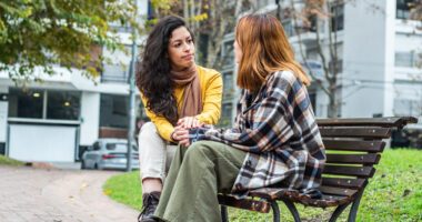 Two young women sit on a city park bench, talking to each other and actively listening.