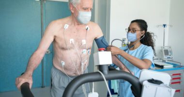 Senior man on treadmill, hooked up to EKG wires as provider tests his blood pressure with an arm cuff
