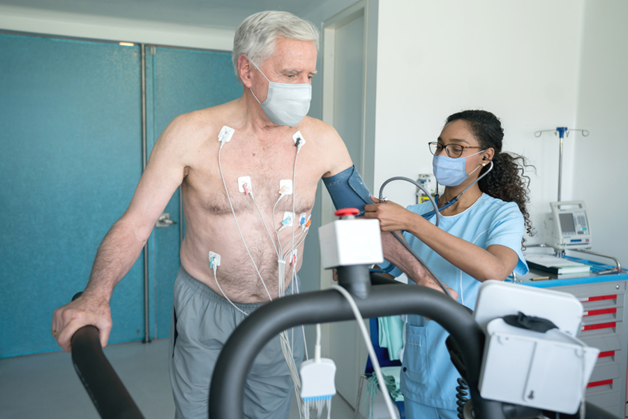 Senior man on treadmill, hooked up to EKG wires as provider tests his blood pressure with an arm cuff