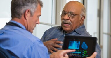 Older man talking to provider and touching chest, while his provider looks at an image of a heart on a tablet