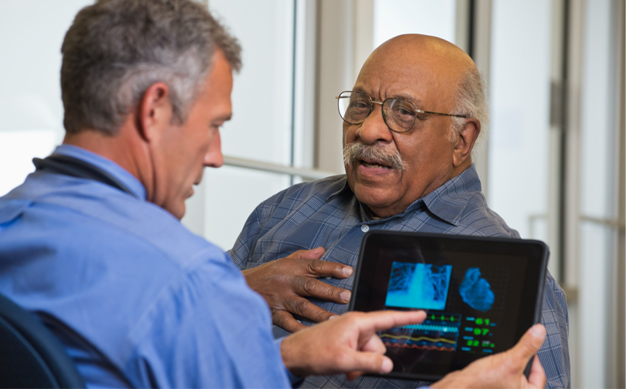 Older man talking to provider and touching chest, while his provider looks at an image of a heart on a tablet