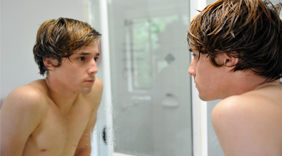teenage boy looks at his reflection in a bathroom mirror