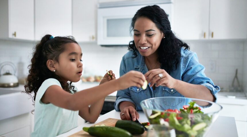Mom and elementary-aged daughter work together to prepare a salad in a brightly-lit kitchen