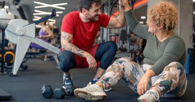 A personal trainer high-fives his client in a gym. Both wear fitness clothes and free weights are on the floor.