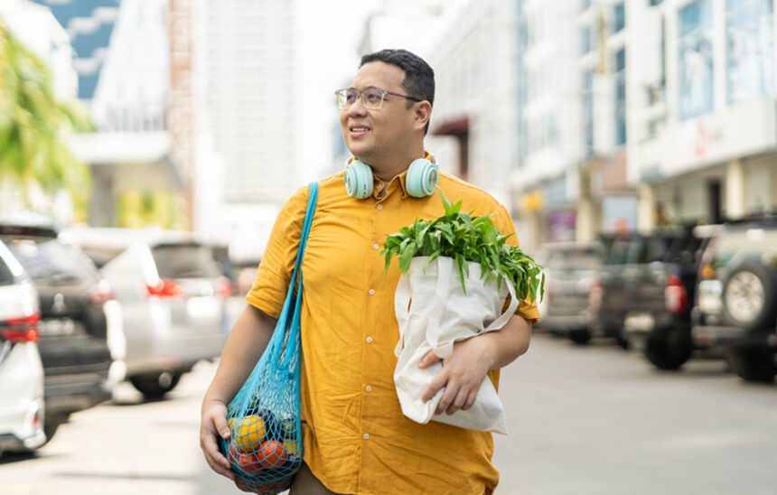 Man walks happily down city street, holding re-usable bags filled with vegetables, presumably from farmer's market