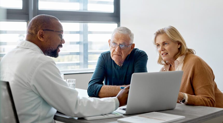 Older couple sits at a desk with provider, in discussion while the provider points to a laptop screen