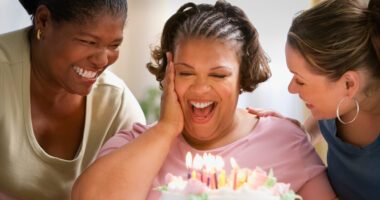 Obese woman smiles at the candles on her birthday cake as two female friends on either side of her celebrate her