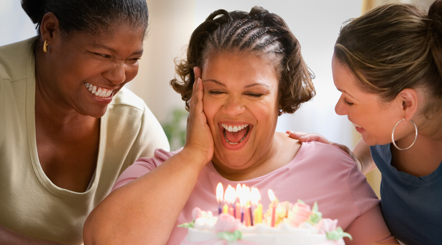 Obese woman smiles at the candles on her birthday cake as two female friends on either side of her celebrate her