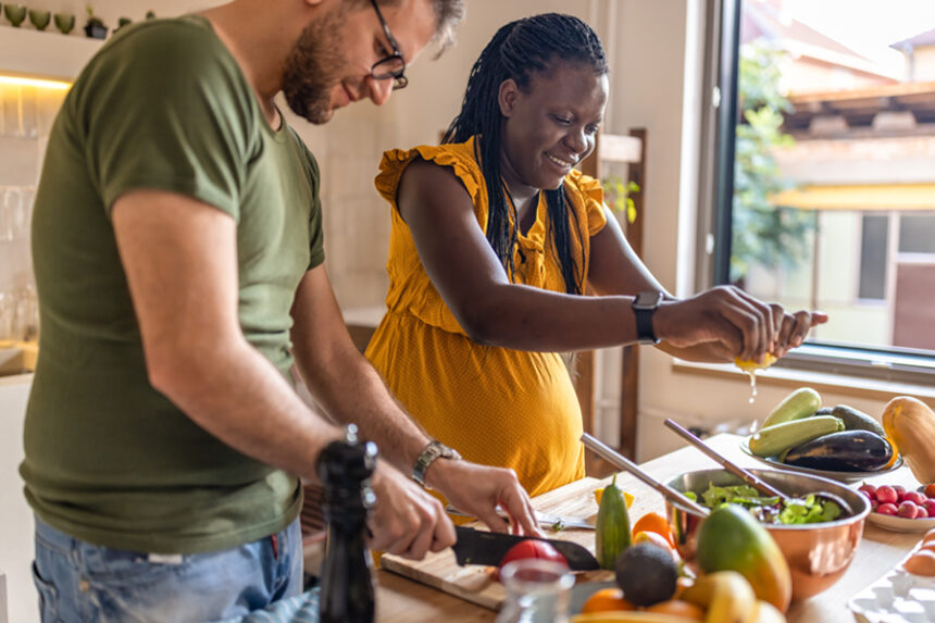 Pregnant woman and her partner prepare vegetables for a meal in a sunny kitchen
