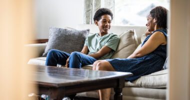 Teenaged boy sits on couch, smiling and talking to his mom
