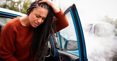 Woman clutches her head in pain, sitting in the passenger seat of a car with the door open as she is stepping out of the car, as if just having been in a car accident