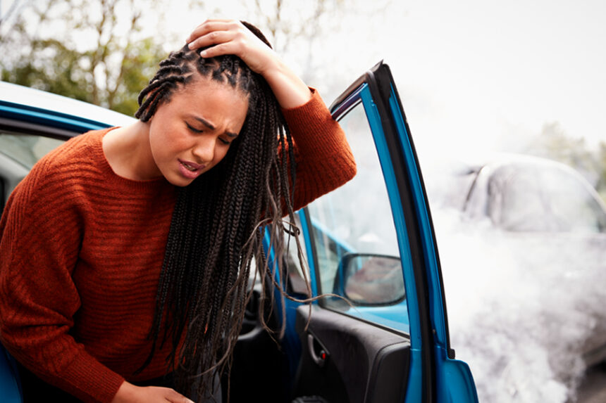 Woman clutches her head in pain, sitting in the passenger seat of a car with the door open as she is stepping out of the car, as if just having been in a car accident