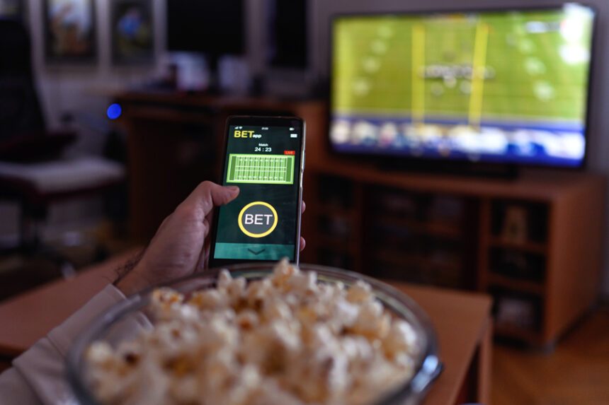 Close view of a smartphone open to a sports betting app while a football game is on the TV in the background