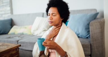 Woman holds throat in pain while holding a cup of tea and wearing a blanket over her shoulders