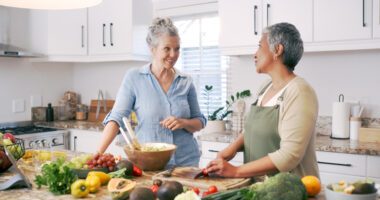 Two menopause-aged women talk in a brightly-lit kitchen while they prepare a salad