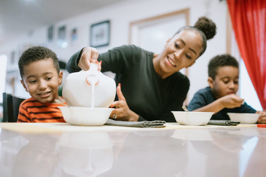 Mom pours milk into a cereal bowl for your young song, while another young son eats his cereal next to her