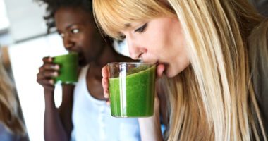 Two women each drink green smoothies from a glass.