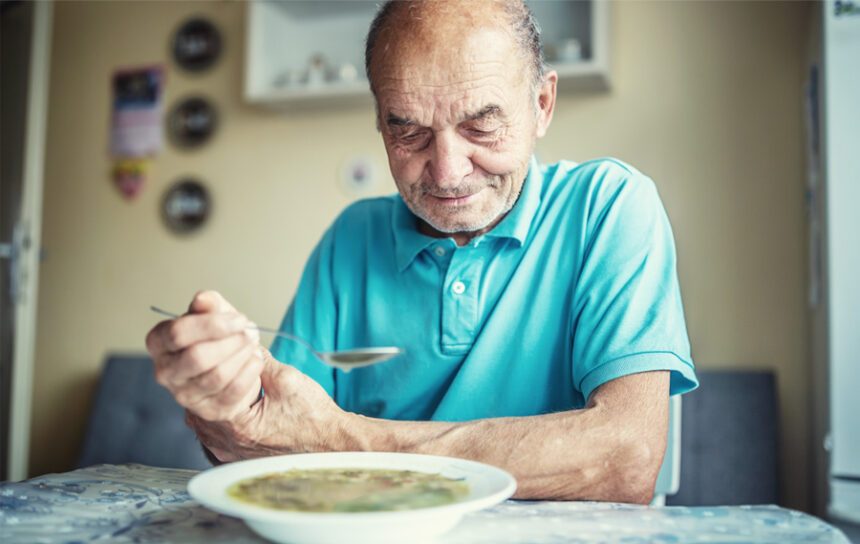 senior man eats a bowl of soup sitting at a small kitchen table