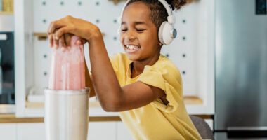 elementary-aged girl makes a fruit smoothie in a blender while wearing headphones