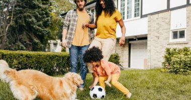 Mom, dad, and son play soccer with a golden retriever in a front yard with a house in the background