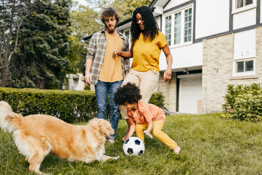 Mom, dad, and son play soccer with a golden retriever in a front yard with a house in the background