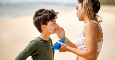 A mom puts sunscreen on her son's face while they stand at the beach