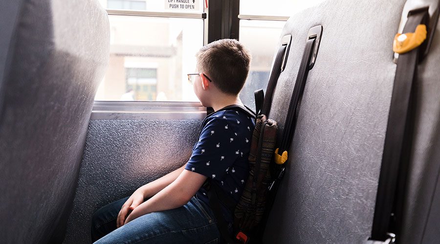 elementary-aged boy sits on school bus in a seat by himself, looking out of the window