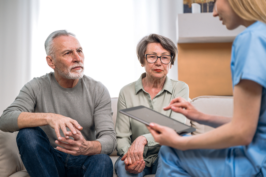 Older man and his wife sit on a couch, speaking to a provider in blue scrubs