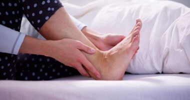 close-up image of adult woman sitting on a bed, clutching ankle and foot