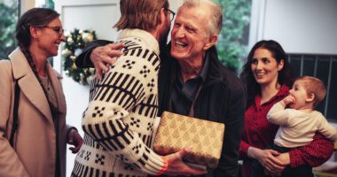 Adult son and father hug, both wearing holiday winter sweaters and holding gifts