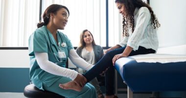 provider examines the ankle of a pediatric patient sitting on exam table while mother watches in background