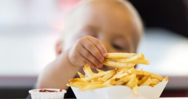 a baby reaches for a french fry in a pile of fries served in a white dish, a paper cup of ketchup on the side