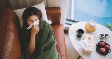 woman lies on a couch, covered in a blanket and blowing her nose with a tissue