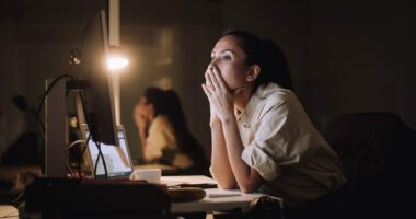 Woman sits at desk in a dark room, hands over her mouth in pensive, frustrated look