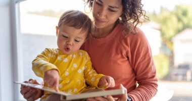 Toddler sits on mom's lap, both looking at a board book together