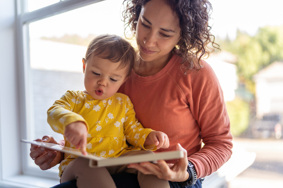 Toddler sits on mom's lap, both looking at a board book together