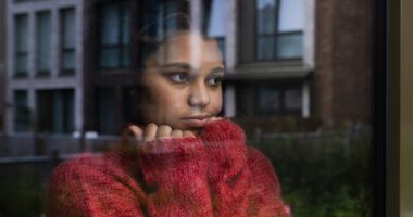 young woman wearing a an oversized sweater looks out a window with a melancholy look on her face