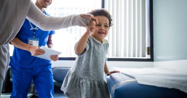 A little girl climbs happily onto an exam table with help from mom's hand while a provider smiles in the background