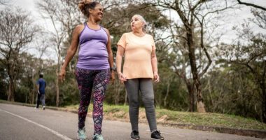 Two middle-aged women wearing workout clothes are walking side by side on a walking path