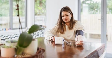 Woman sits at her kitchen table, using an at-home blood pressure monitor.