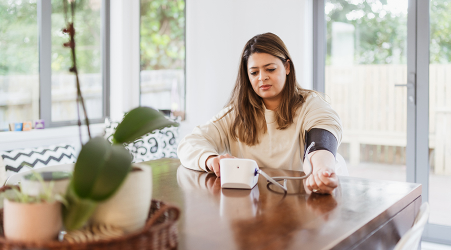 Woman sits at her kitchen table, using an at-home blood pressure monitor.