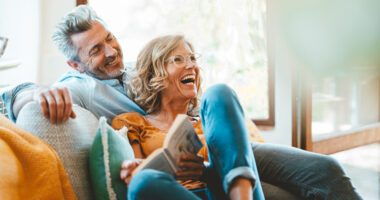 menopause-aged woman holds a book, laughs with male companion as they sit relaxingly on a couch