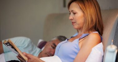 menopause-aged woman sits in bed reading, while her husband sleeps next to her