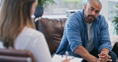 Man sits on couch talking while a therapist sits in a chair, taking notes